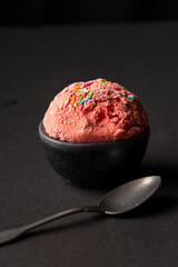 Top view of scoop of strawberry ice cream in black bowl with spoon, selective focus, black background, vertical, with copy space