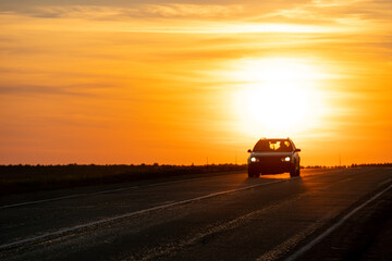 Fototapeta premium The silhouette of a car on the road against the background of the sun. The car is driving on the highway during sunset. The concept of travel and freedom.
