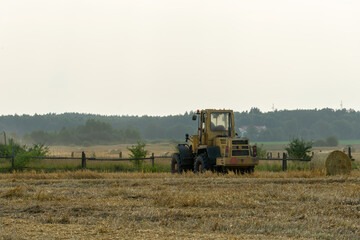 A yellow tractor with the help of a manipulator puts round bales of hay on a trailer....