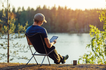Man is reading an e-book on the shore of a forest lake at sunset in the evening.