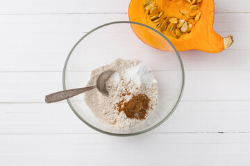 Glass bowl with oatmeal flour, baking powder and ground cinnamon, half a pumpkin with seeds on a white wooden background, top view. Stage of cooking delicious vegan dessert