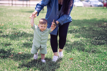 Cute baby boy learning to walk with careful mother help on lush lawn grass in spring park family of...