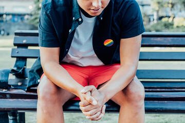 Cropped no face sad frustrated LGBTQ person with rainbow badge on t-shirt sitting on park bench...