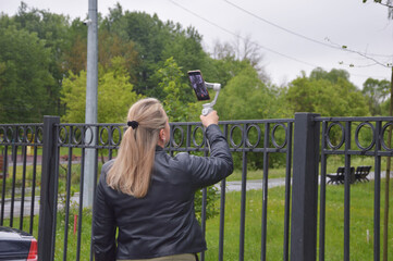 Fototapeta na wymiar A blonde blogger girl records a video while holding a phone on a selfie stick with a stabilizer in a city park.