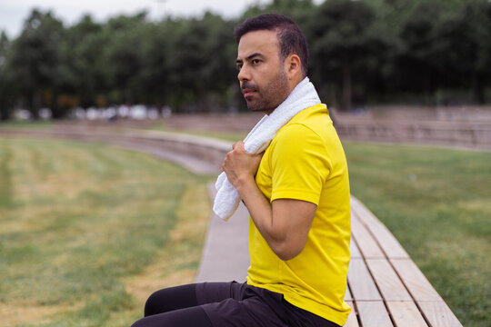 Seated on a wooden bench in the lush green city park, a handsome man in a tracksuit takes a moment to rest during his routine training on the outdoor sports ground.
