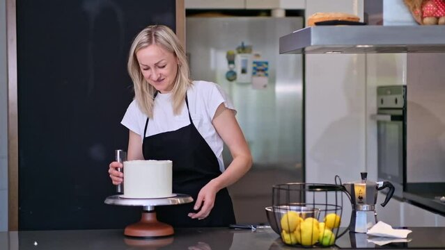 Happy blonde woman finishing processing white mastic on sugar free cake with pastry scraper self-made confectioner enjoying preparing custom confectionery for anniversary