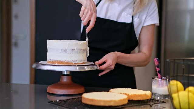 Food blogger confectioner preparing delicious lactose free cake for event at home woman in black apron standing at kitchen table demonstrates smearing white cream on handmade confectionery