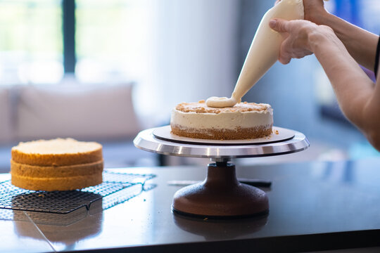 Woman making handmade cake putting fresh white cream on layer with pastry cone skilled housewife standing at kitchen table preparing delicious handmade custom pastry at home closeup