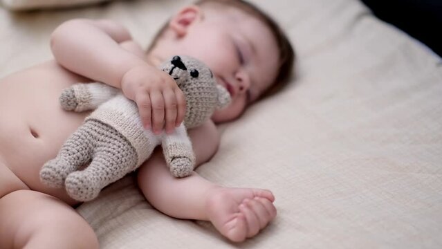 Cute chubby baby sleeping on back holding knitted bear toy with hand sweet little boy with closed eyes taking nap in comfortable bed in nursery at home