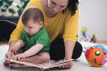 Fototapeta premium Chubby baby boy examining book with colorful pictures sitting near mother on white rug near toys woman educating little son at home developing new skills
