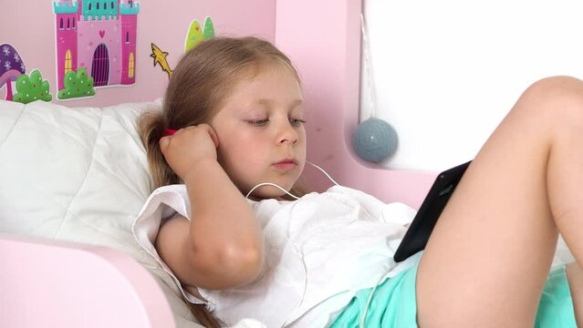 girl lies with the phone. Close-up of a cute caucasian little girl lying on a bed in a children's room wearing headphones and watching a movie. The child poses with a smartphone. telephone addiction