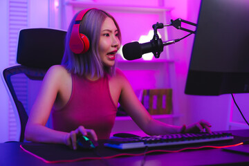 Young cute woman talking to players and audience on computer in neon led light room at home, Excited smiling gamer Asian girl waring headset with a mic playing an online video game live streaming