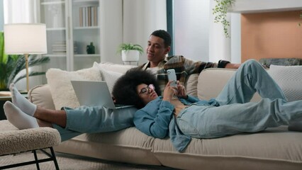 African American happy family relaxing at new home on couch couple on sofa man husband freelancer boyfriend working with laptop computer woman wife girlfriend scrolling mobile phone gadget addiction - Powered by Adobe