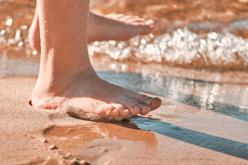 Children's feet stand on the sand of the sea beach and are washed by the waves, close-up. A child of 10-12 years old plays near the water in summer.