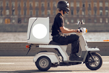 The courier on a white electric scooter is in a hurry to complete the order. A man carries a delivery on a moped with a trunk along the river embankment. Courier service delivering the parcel.