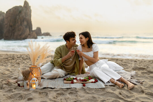 Loving romantic couple having picnic on sandy beach near ocean, drinking wine and enjoying date at coastline, copy space - Powered by Adobe