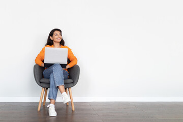 Positive young eastern woman sitting in armchair, using laptop