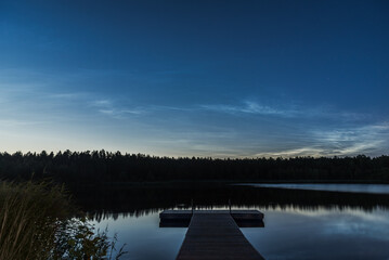 Noctilucent clouds over the forest lake in Latvia at July night. Wooden pier on foreground.