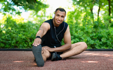 Positive attractive african american athlete man in sportswear and headphones stretching legs