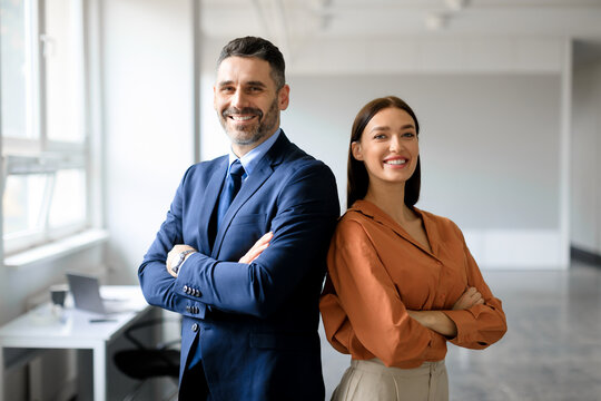 Successful businessman and businesswoman standing back to back with arms crossed and smiling at camera, office interior