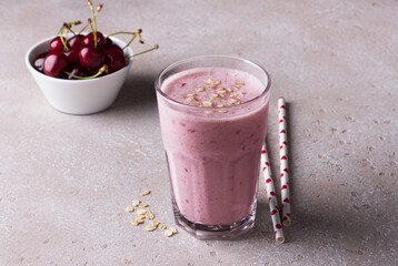 Milkshake with cherries, yogurt and oatmeal, drinking straws, bowl with fresh cherries on a beige textured background