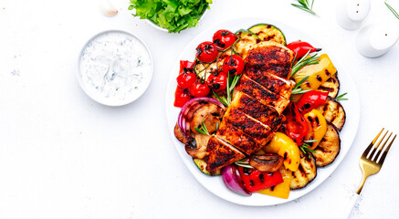 Grilled chicken breast and various vegetables. Colorful paprika, zucchini, eggplant, mushrooms, tomatoes, onion with rosemary on plate, white table background, top view