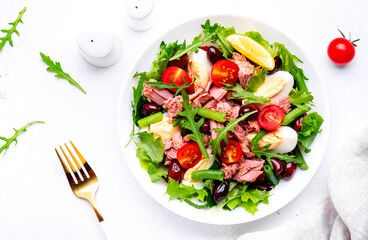 Nicoise salad with canned tuna, cherry tomatoes, boiled eggs, green beans and red olives on plate, white table background, top view