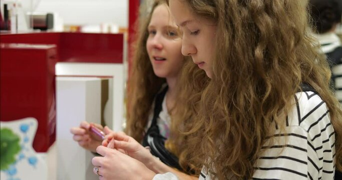 Two teen girl siblings stand at make-up showcase counter and hold lip gloss. Teenage sisters are shopping for cosmetics in beauty products supermarket department.