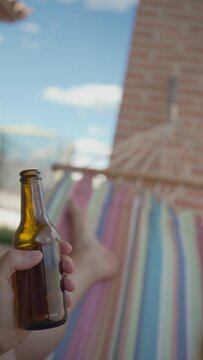 Unrecognizable person relaxing with a beer on his hammock. Point of view (vertical) video of an anonymous young man lying on his backyard hammock swinging while holding a fresh beer in a bottle.