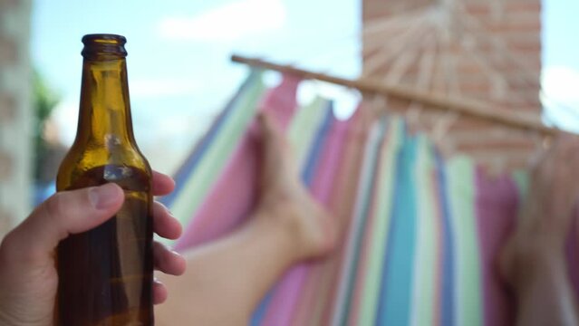 Unrecognizable person relaxing with a beer on his hammock. Point of view video of an anonymous young man lying on his backyard hammock swinging while holding a fresh beer in a bottle.