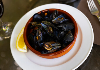 Fototapeta premium Appetizing steamed mussels in shells served with garlic butter sauce and lemon wedge in clay bowl. Mediterranean seafood appetizer..