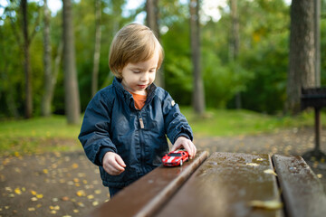 Cute toddler boy playing with red toy car outdoors. Kid exploring nature. Small child having fun with toys. Autumn activities for children.