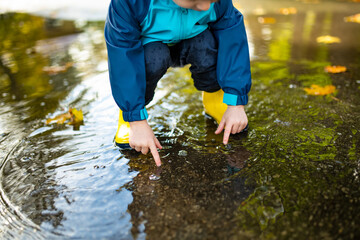 Adorable toddler boy wearing yellow rubber boots playing in a a puddle on sunny autumn day in city park. Child exploring nature. Fun autumn activities for kids.