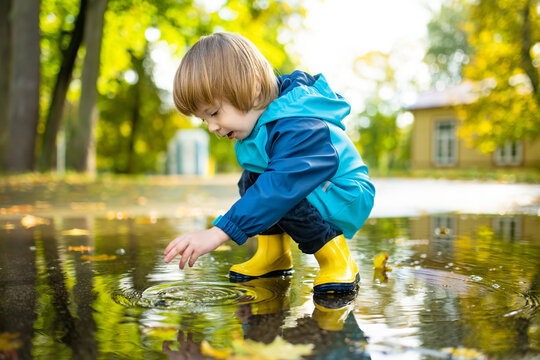 Adorable toddler boy wearing yellow rubber boots playing in a a puddle on sunny autumn day in city park. Child exploring nature. Fun autumn activities for kids.