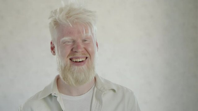 Portrait of young laughing bearded man with albinism, wearing white clothes on white background. Medium close up of albinos. Man with unusual appearance. High quality 4k footage