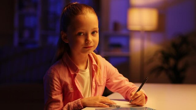 Portrait of pretty little girl sits at home in the evening writting in his notebook, looking at the camera, blinking and smile. Close up, detailed shot, full face.