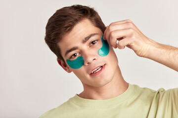 A millennial guy with brown hair practicing skincare with under eye hydrogel patches in a studio. Young smiling caucasian man applying cosmetic patches on his face. Men's self-care.