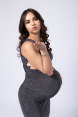 Elegance caucasian pregnant 30 year old brunette woman with long hair in a shiny tight dress, Sending an air kiss. Studio white background. Concept 9 months pregnancy, aesthetic pregnancy, beauty.