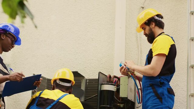 Adept repairmen diverse team working with manifold gauges to check air conditioner refrigerant tank in need of maintenance. Professional workers using barometer benchmarking hvac system component