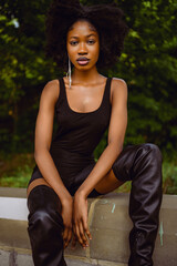 Fashion outdoor street style portrait Beautiful young African American woman posing outside on urban city landscape summer day wearing total black high leather boots.