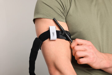Soldier in military uniform applying medical tourniquet on arm against light grey background,...