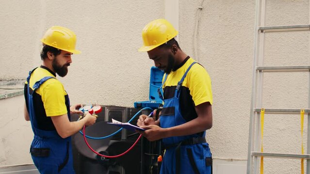 Adept repairmen working with manifold meters to check air conditioner refrigerant levels, writing result on clipboard. Proficient workers using barometer benchmarking outdoor hvac system tool