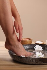 Obraz na płótnie Canvas Woman soaking her foot in bowl with water, spa stones and chrysanthemum flowers on wooden surface, closeup. Pedicure procedure