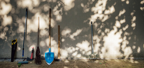 Tools for the care of the garden and the house stand near the gray wall in the shade of trees and...