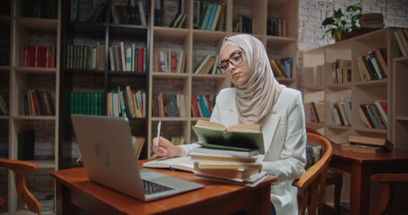 Female muslim student is studying at desk in library, using laptop and books. Girl wearing hijab is preparing for exams - student lifestyle, modern Islam concept close up
