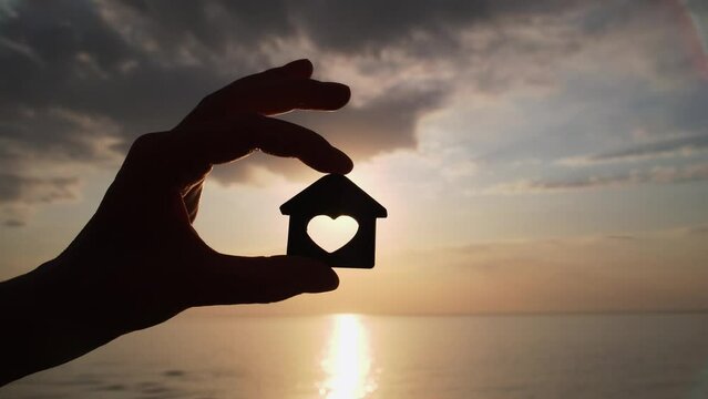 Close-up shot of woman's hand holding wooden house model on sunset and sea background. Seaside dream house concept. Choosing place for a new home.