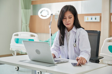 Young beautiful Asian female doctor online working with laptop and smartphone on medical consultation application via internet at working desk in hospital clinic.