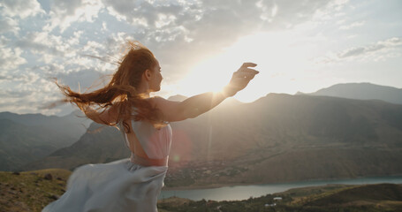 Woman in white dress standing on top of a mountain with raised hands while wind is blowing her dress and red hair - freedom, nature concept