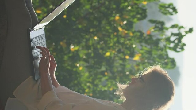 Vertical shot of blonde woman typing on a laptop keyboard in sunshine outdoor. Female writer working on the rooftop with a beautiful view at sunrise.