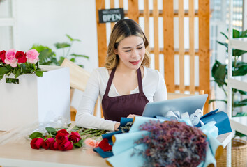Asian happy cheerful female florist designer shop owner checking smelling quality of red roses blooming in store while using laptop notebook computer calculating finanicial target in floral studio